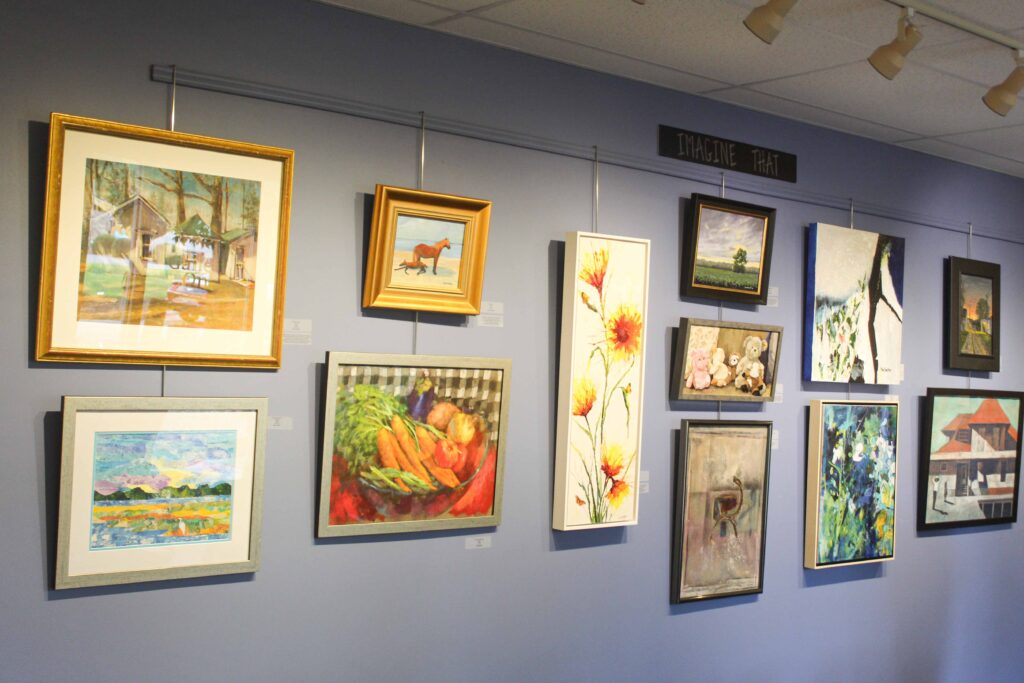 Bethany Boardwalk Arts Festival A light purple wall displays various framed paintings, including landscapes, flowers, animals, and abstract art. A sign above reads IMAGINE THAT. Ceiling tiles and lights are visible above the paintings.