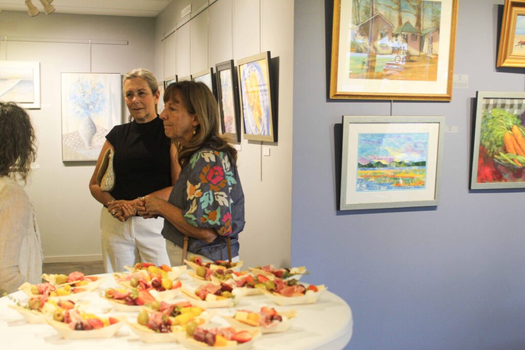 Bethany Boardwalk Arts Festival Three women converse in an art gallery filled with colorful paintings. A round table in the foreground holds plates of assorted snacks, including fruit and cheese. The atmosphere appears social and inviting.
