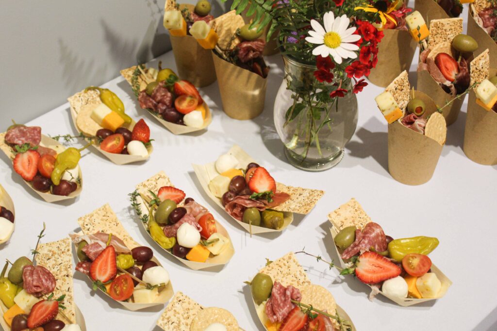 Bethany Boardwalk Arts Festival Small paper cups and boats filled with cheese, salami, strawberries, olives, and crackers are arranged on a white tablecloth. A glass vase with colorful flowers is placed among the snacks.