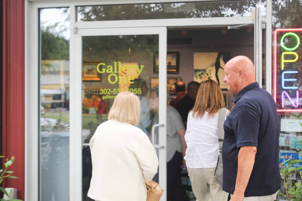 Bethany Boardwalk Arts Festival Several people enter Gallery One through a glass door. The OPEN sign is lit, and artwork is visible inside. A man in a navy shirt stands outside, while others head into the brightly-lit space.