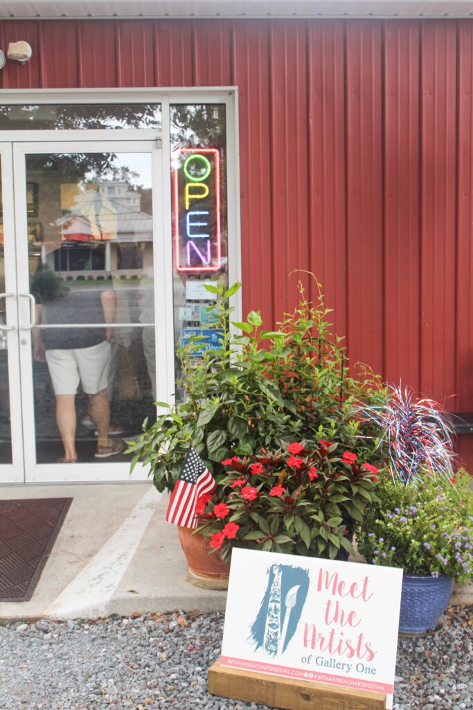 Bethany Boardwalk Arts Festival A person stands by a glass door under a neon OPEN sign. Outside, theres a pot of flowers with an American flag and a sign that reads, Meet the Artists of Gallery One..