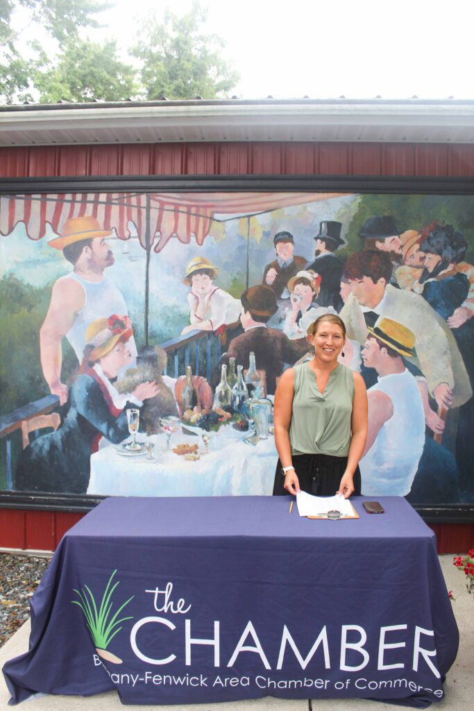 Bethany Boardwalk Arts Festival A smiling woman stands behind a table with a Chamber tablecloth, holding a pen. Behind her is a mural of people dining outdoors. The tablecloth reads Bethany-Fenwick Area Chamber of Commerce..