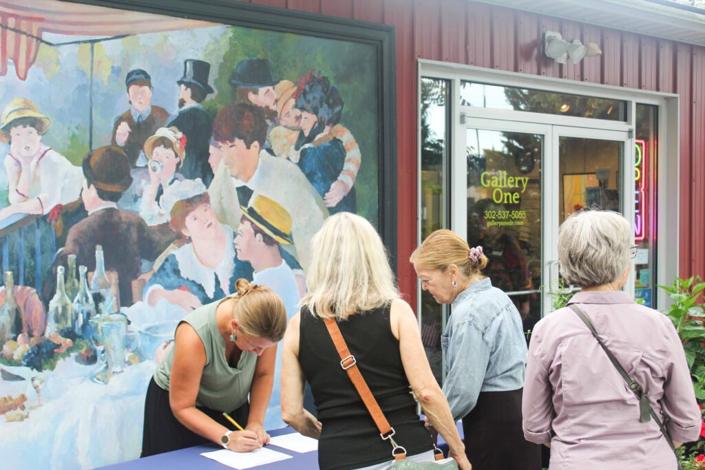 Bethany Boardwalk Arts Festival Four women stand at a table outside an art gallery, one signing a paper. A large mural of people dining outdoors is painted on the wall behind them.