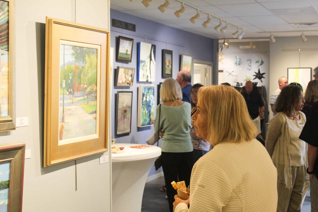 Bethany Boardwalk Arts Festival Visitors at an art gallery walk through a room filled with various paintings on the walls. In the foreground, a woman holds a snack while looking at a framed landscape painting. The gallery is well-lit with track lighting overhead.