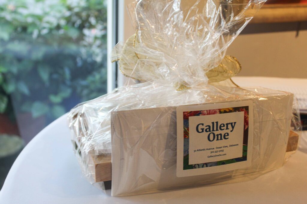 Bethany Boardwalk Arts Festival A gift basket wrapped in clear cellophane with a gold bow sits on a white table. A rectangular sticker on the front reads Gallery One with address and contact details for the Ocean View, Delaware location.