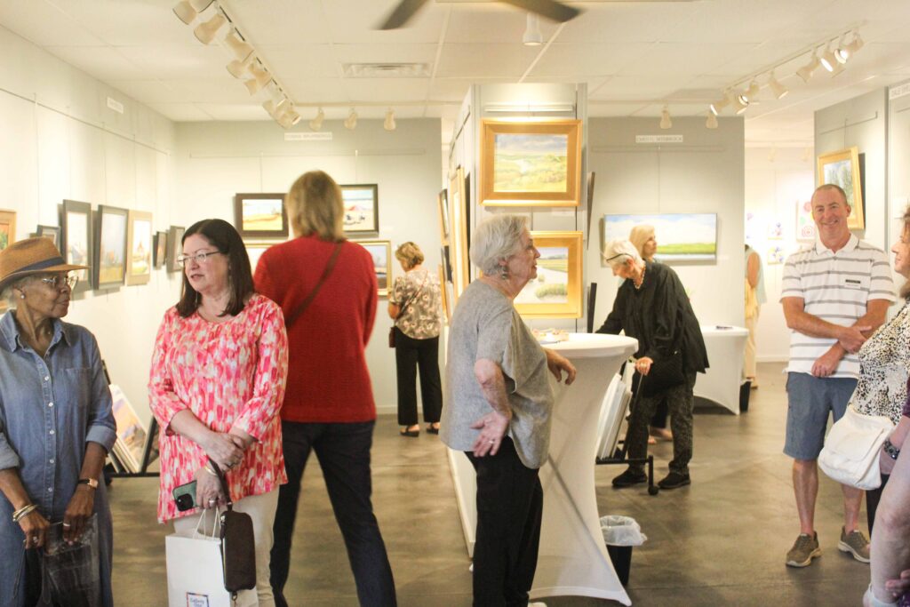 Bethany Boardwalk Arts Festival People view artwork in a brightly lit gallery, chatting and standing near paintings on the walls. Some stand at a tall white table. The atmosphere is casual and social.