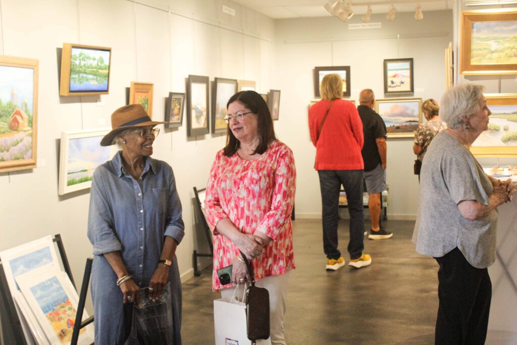 Bethany Boardwalk Arts Festival People view paintings in an art gallery. Two women in the foreground chat and smile, while others admire the artwork on the walls in the background.