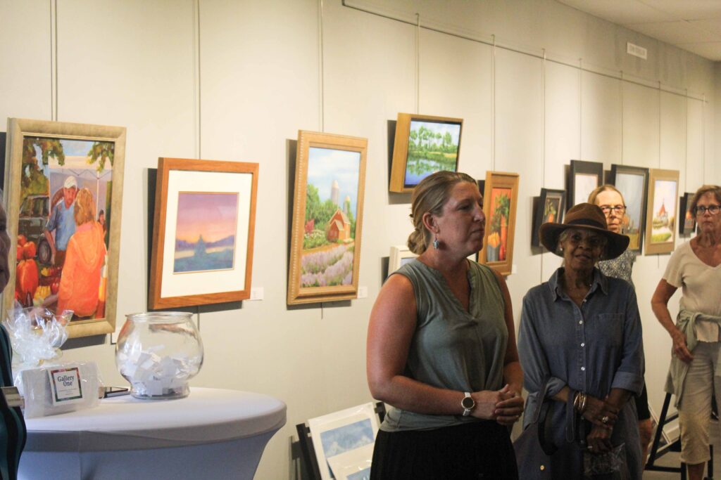 Bethany Boardwalk Arts Festival A woman speaks in an art gallery with landscape paintings on the wall behind her. Three other women stand nearby, listening. A table with a glass bowl and a wrapped item is in the foreground.